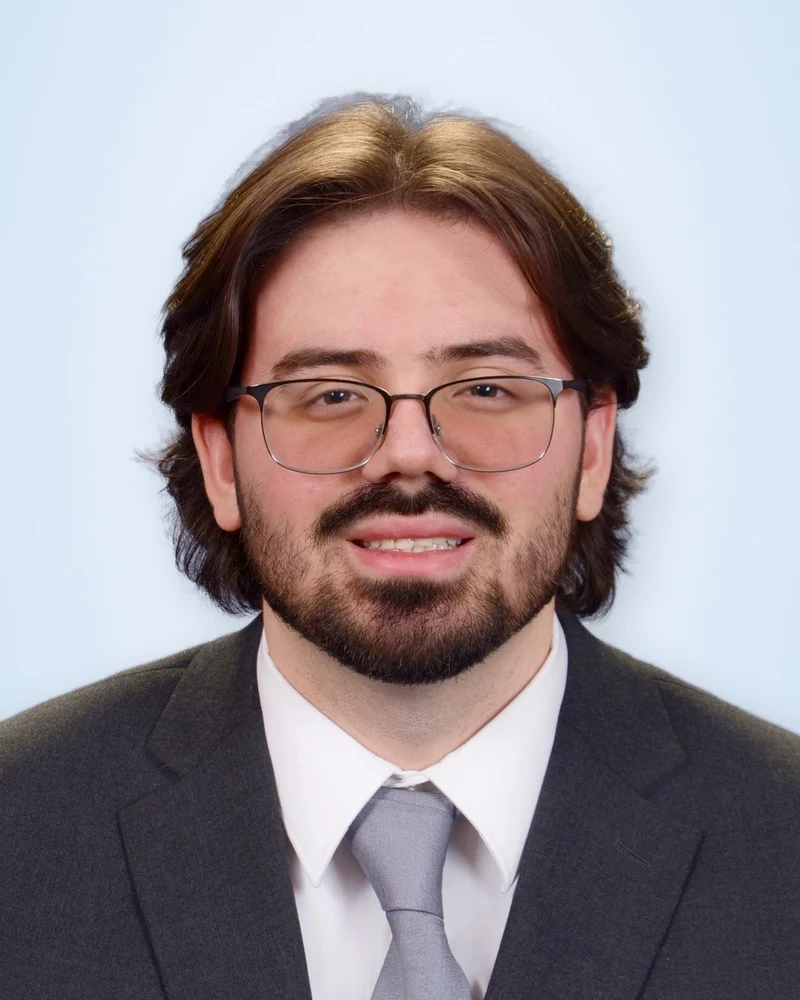 Luke Bratic, smiling, wearing glasses, a dark suit, and a gray tie.